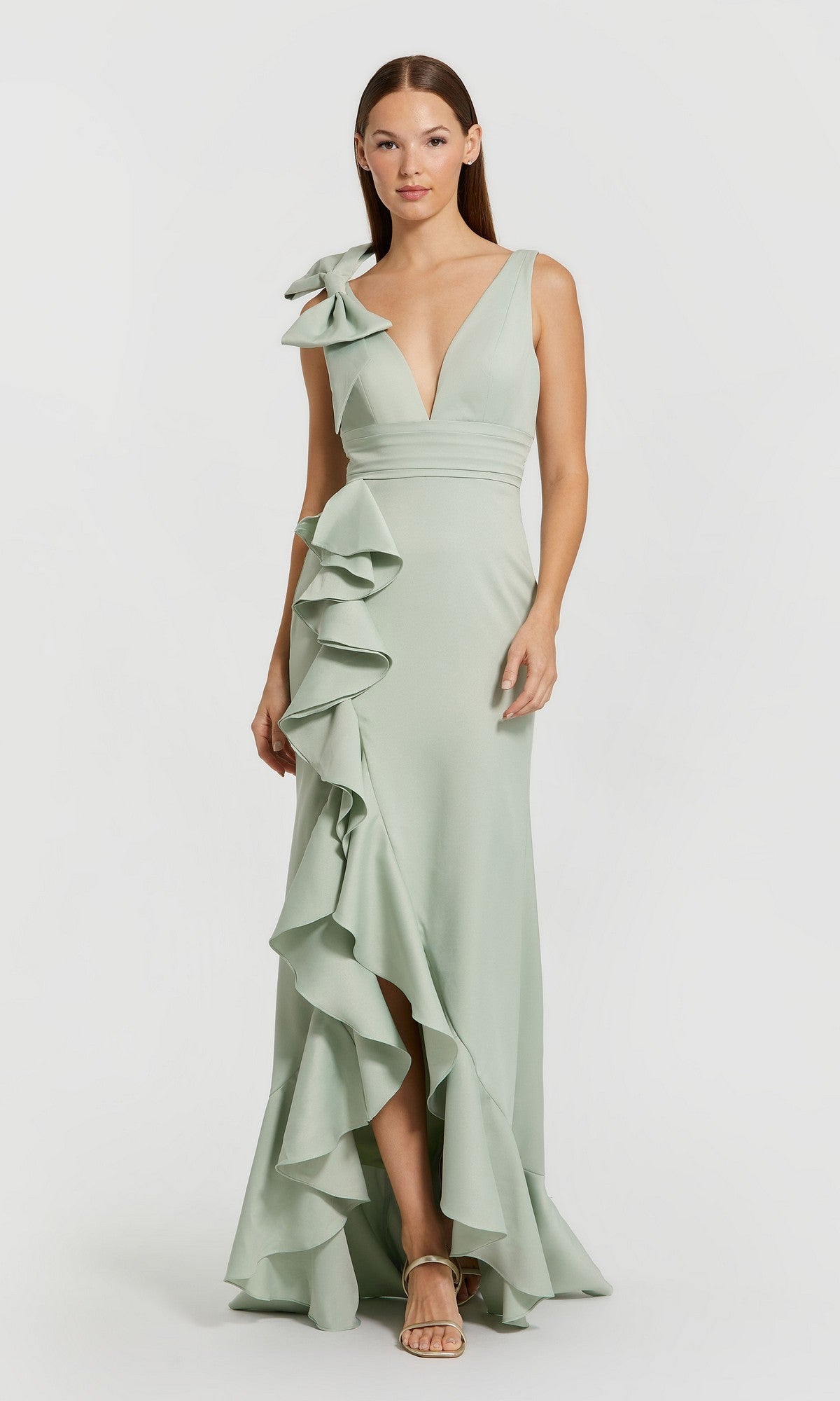 A woman wearing the Mac Duggal 49965 long formal dress in sage—a sleeveless crepe trumpet gown featuring ruffle details on the left side and a bow on the shoulder—standing against a plain background.